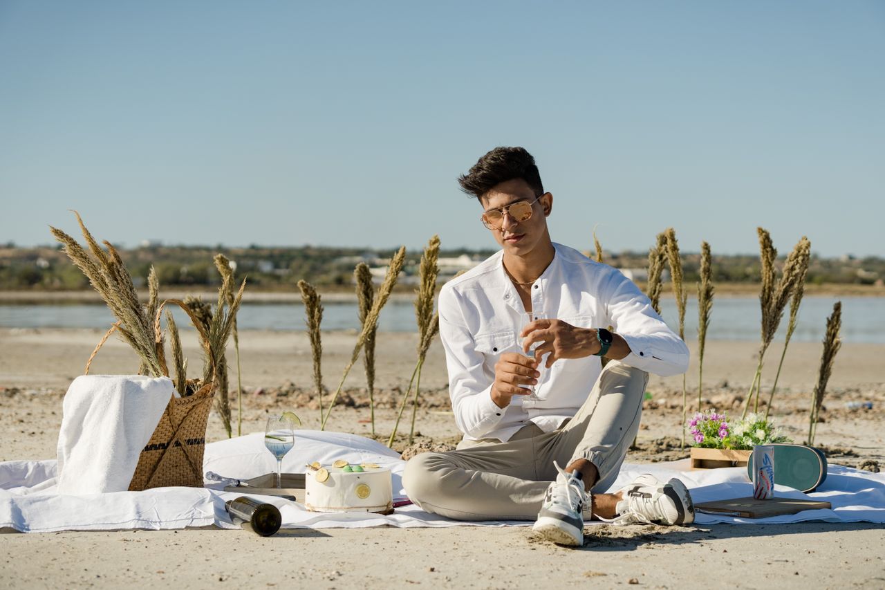 Young man enjoying a sunny beach picnic with stylish sunglasses and casual attire.