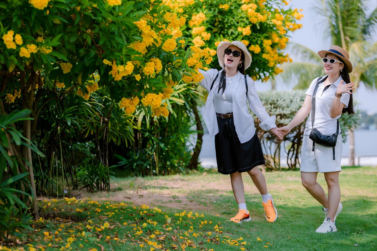 Two women joyfully pose among vibrant yellow flowers in a sunny outdoor garden.